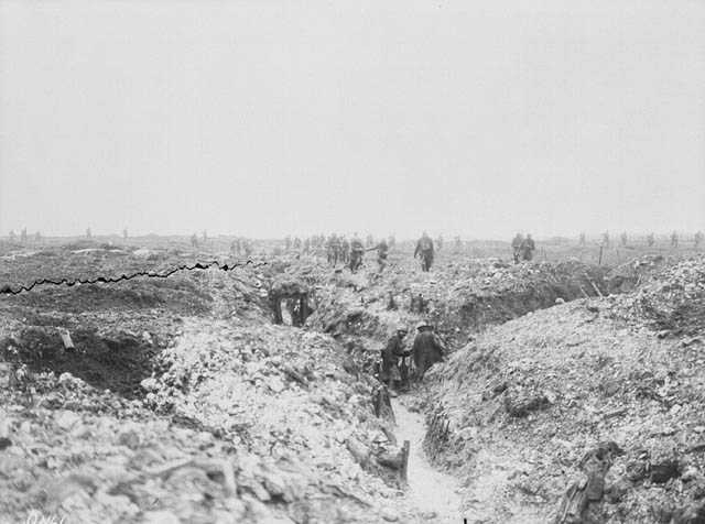 Photo en noir et blanc d’un grand champ boueux avec une tranchée en forme d’Y. Des hommes au loin traversent les tranchées et observent l’intérieur des redoutes. Au-delà des tranchées, d'autres hommes avancent à travers le paysage.